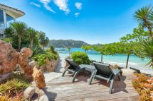 Beachfront wooden deck with reclining lounge chairs, rocky landscaping, tropical plants, and turquoise sea under a clear blue sky.
