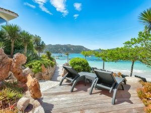 Beachfront wooden deck with reclining lounge chairs, rocky landscaping, tropical plants, and turquoise sea under a clear blue sky.