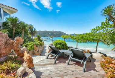 Beachfront wooden deck with reclining lounge chairs, rocky landscaping, tropical plants, and turquoise sea under a clear blue sky.
