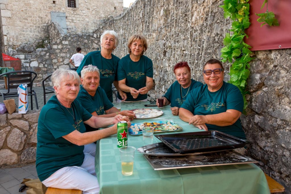 Six people in teal shirts sit around a stone-walled courtyard table, sharing snacks and drinks outdoors.