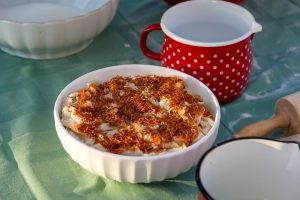 White ceramic bowl filled with creamy coleslaw topped with orange crispy bits on a green table, with a red polka-dotted pitcher in the background.