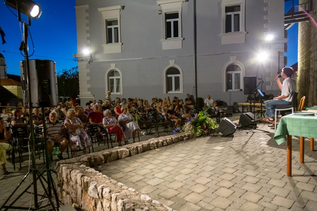 Outdoor evening concert in a town square with a singer on a small stage and a seated audience in front of a white building under blue dusk sky.