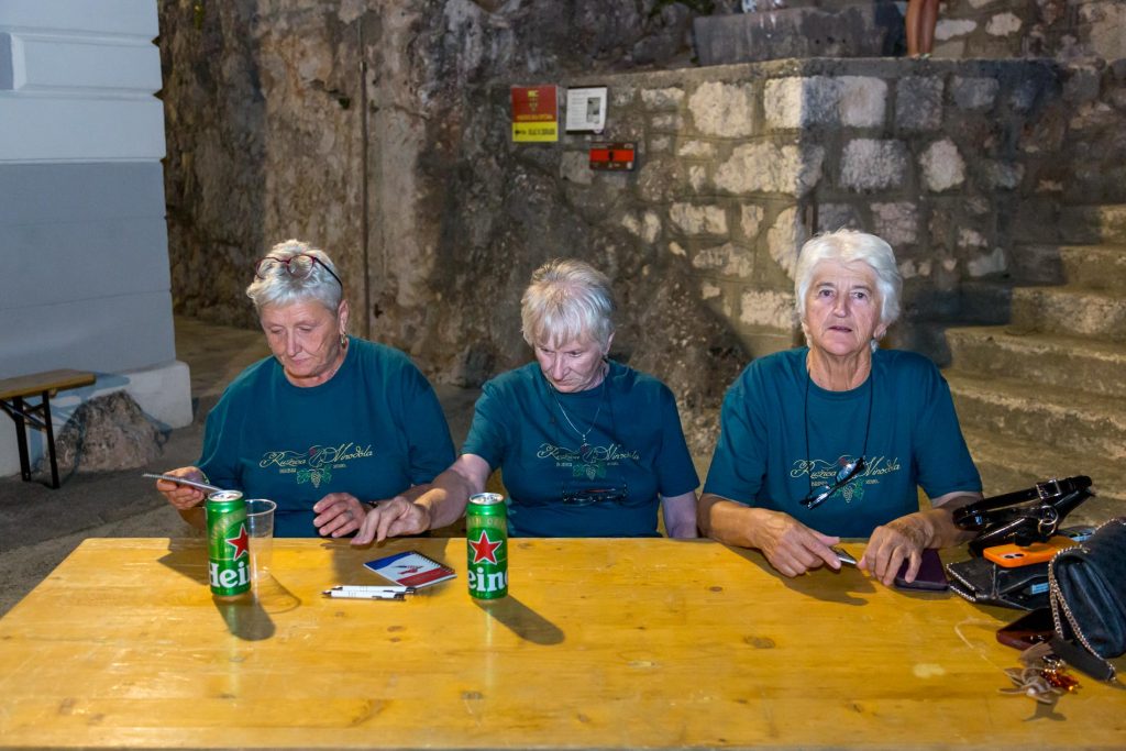 Three older women in teal shirts sit at a wooden table with drinks and papers, against a stone wall backdrop at night.