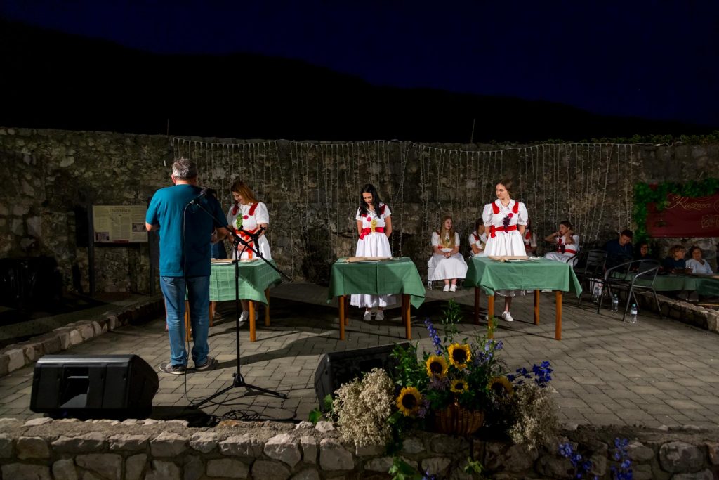 Women in white dresses with red sashes stand behind green-clothed tables on an outdoor stage at night, with string lights and a stone wall behind them and a man at a microphone in the foreground.