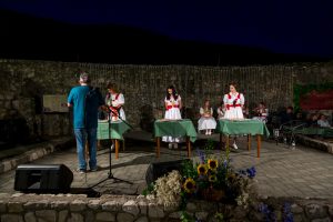Women in white dresses with red sashes stand behind green-clothed tables on an outdoor stage at night, with string lights and a stone wall behind them and a man at a microphone in the foreground.
