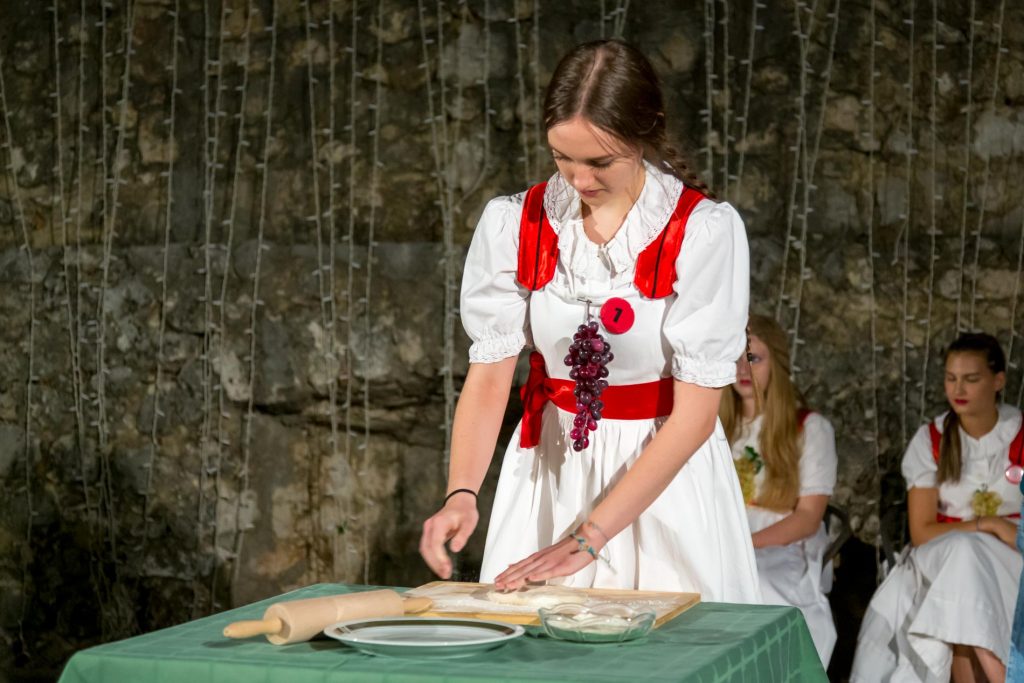 Young woman in a white dress with red sash kneading dough on a green table, rolling pin and plate nearby, others in similar outfits in the background.