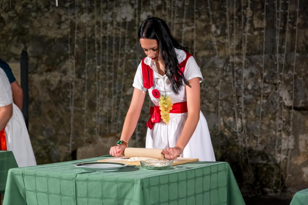 Woman in a white dress with red accents rolls dough with a rolling pin at a table covered in a green cloth, near a stone wall backdrop.