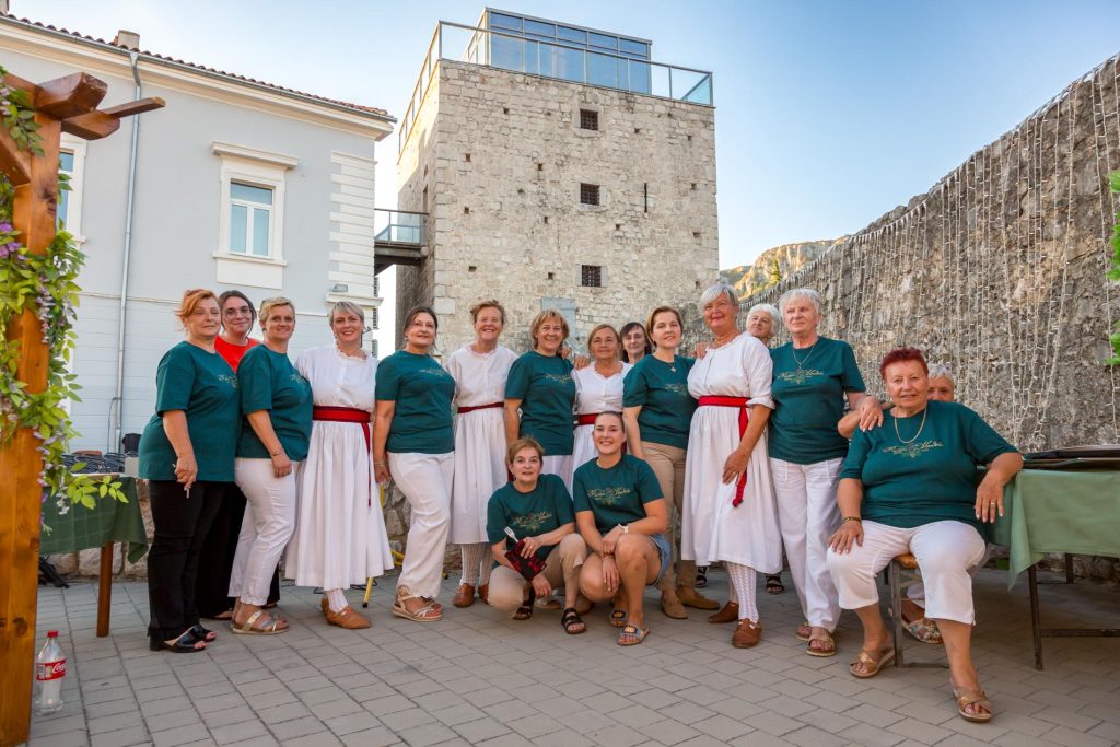 Group of women in teal blouses and white skirts posing together in an outdoor courtyard with a stone tower in the background.