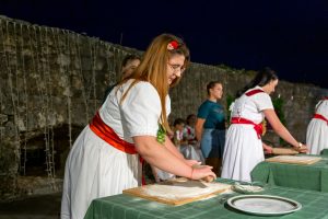 Women in white dresses with red sashes roll dough at an outdoor night event, green-table setup in foreground.