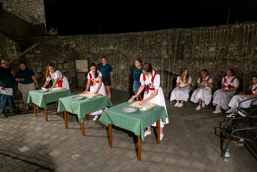 Group of young women in white dresses with red sashes demonstrating dough rolling at outdoor night event, with onlookers against a stone wall and string lights.