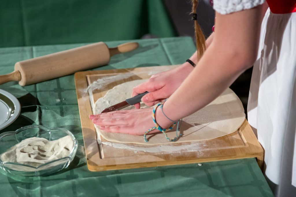 Hands flattening dough on a wooden board with a knife, rolling pin nearby in a kitchen setting.