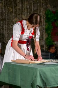 Young woman in white dress with red sash rolling dough on a cutting board with a rolling pin at a table covered in green cloth, grapes hanging from her neck pendant backdrop.