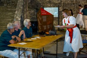 Seniors sitting at a wooden table in a stone venue as a woman in a white dress with a red sash serves a plate to them, smiling.