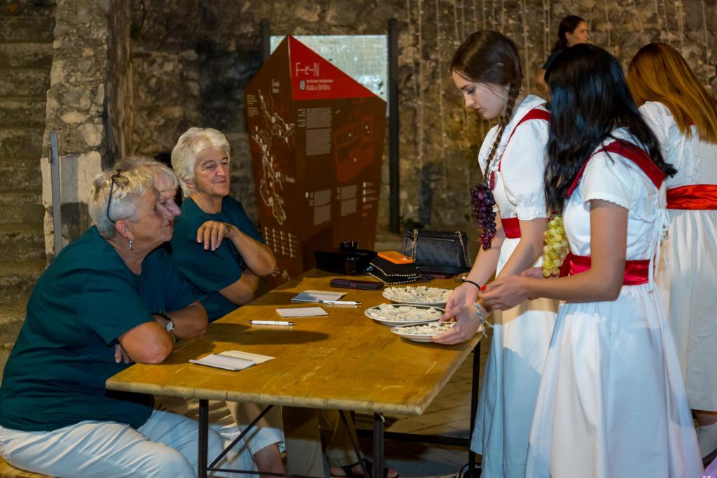 Older adults seated at a wooden table watching two young women in white dresses share small pastries at an event in a stone-walled room.