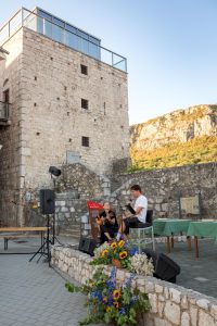 Two musicians playing guitars in a stone courtyard, with a microphone, speakers, and a colorful flower arrangement in the foreground.