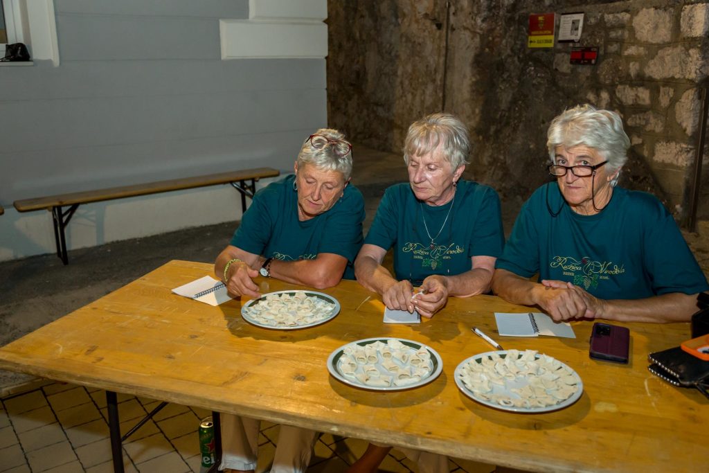 Three elderly women in teal shirts sit at a wooden table, arranging dumplings on plates in a rustic indoor space.