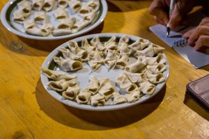Plate of raw tortellini arranged on a wooden table, with a person writing in a notebook in the background.