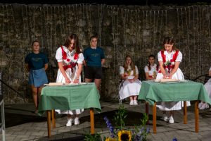 Nighttime outdoor performance: women in white dresses with red accents roll dough at tables covered in green cloth, flanked by watchers in teal shirts against a stone wall with string lights.