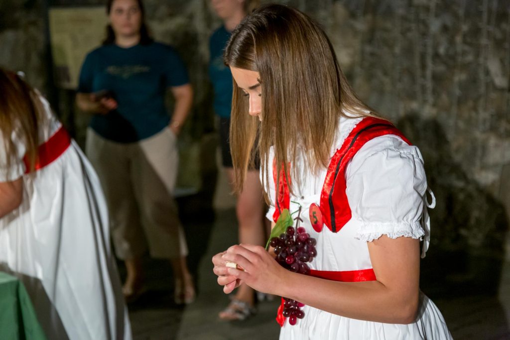 Girl in a white dress with red trim holding a bunch of grapes in a dim indoor setting, with blurred people in the background.