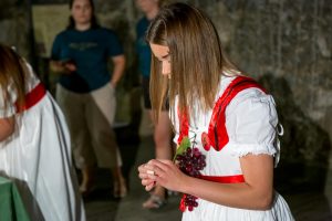 Girl in a white dress with red trim holding a bunch of grapes in a dim indoor setting, with blurred people in the background.