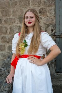 Young woman in a white dress with a red sash, wearing braids and red lipstick, posing outdoors with a grape cluster accessory on her chest.