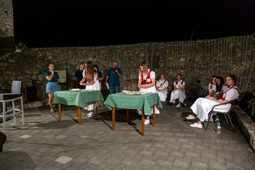 Nighttime outdoor scene with people in traditional white dresses and red sashes around green-tableclothed tables in a stone courtyard.