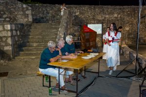 Nighttime outdoor event with three seniors at a wooden table, plates and drinks, as two young women in white dresses with red sashes serve nearby.