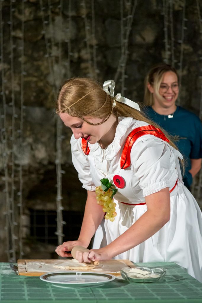 A young woman in a white dress with red trim rolls dough on a wooden board; another smiling woman stands behind her in a dim background.