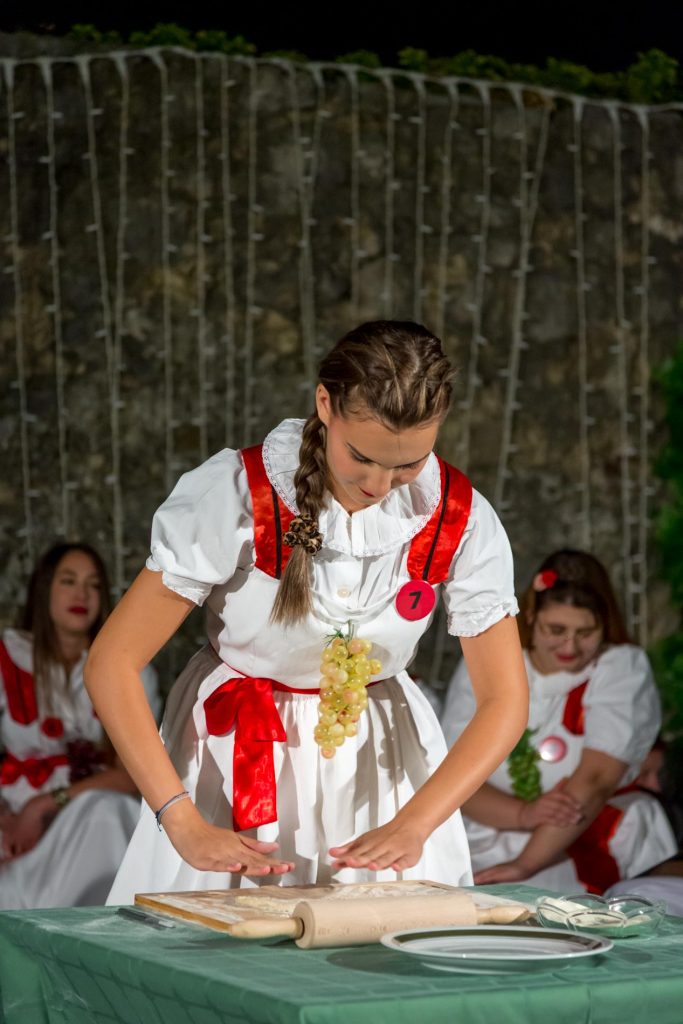 Young woman in a traditional folk dress rolling dough on a green table, with a grape bunch at her waist and others seated in the background.