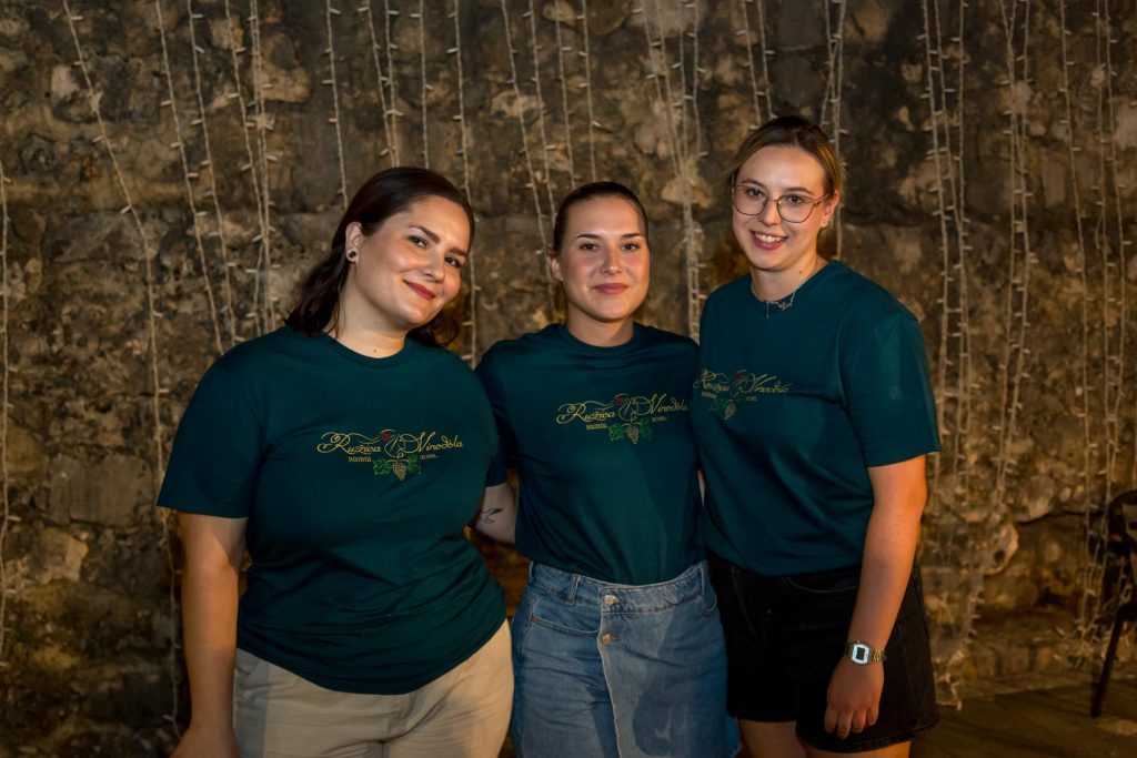 Three women wearing teal T-shirts stand side by side, smiling at the camera in front of a rustic stone wall with vertical string-like decor.