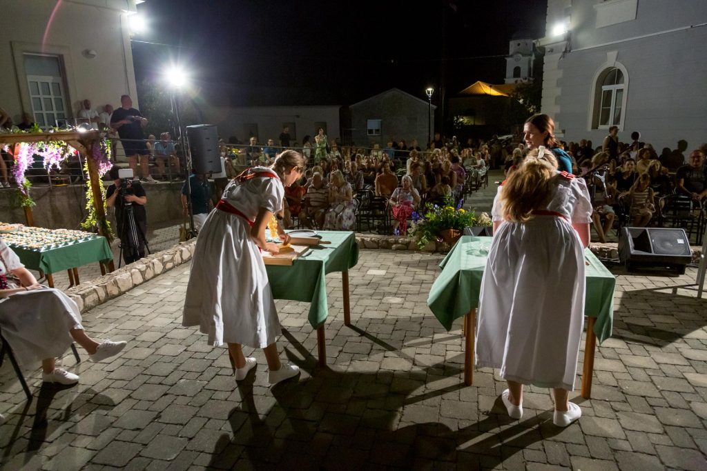 Women in white dresses with red sashes standing at tables cutting or preparing food during an outdoor night event, with a seated crowd in the background and stage lighting.