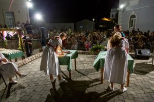 Women in white dresses with red sashes standing at tables cutting or preparing food during an outdoor night event, with a seated crowd in the background and stage lighting.