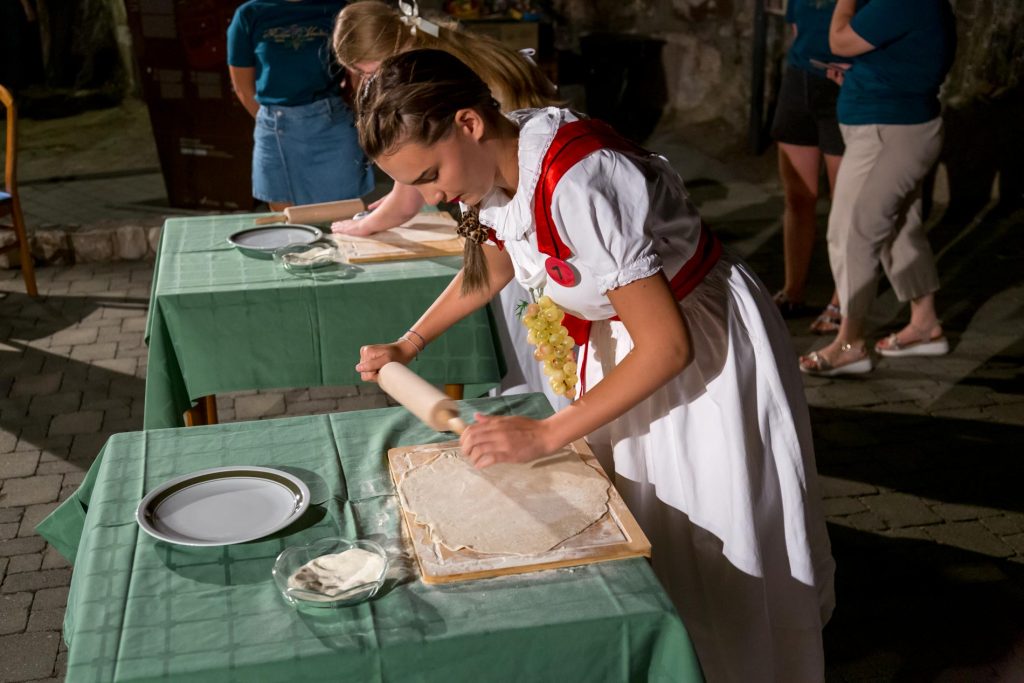 Young woman in a traditional dress rolls dough with a rolling pin at an outdoor event, with onlookers in the background.