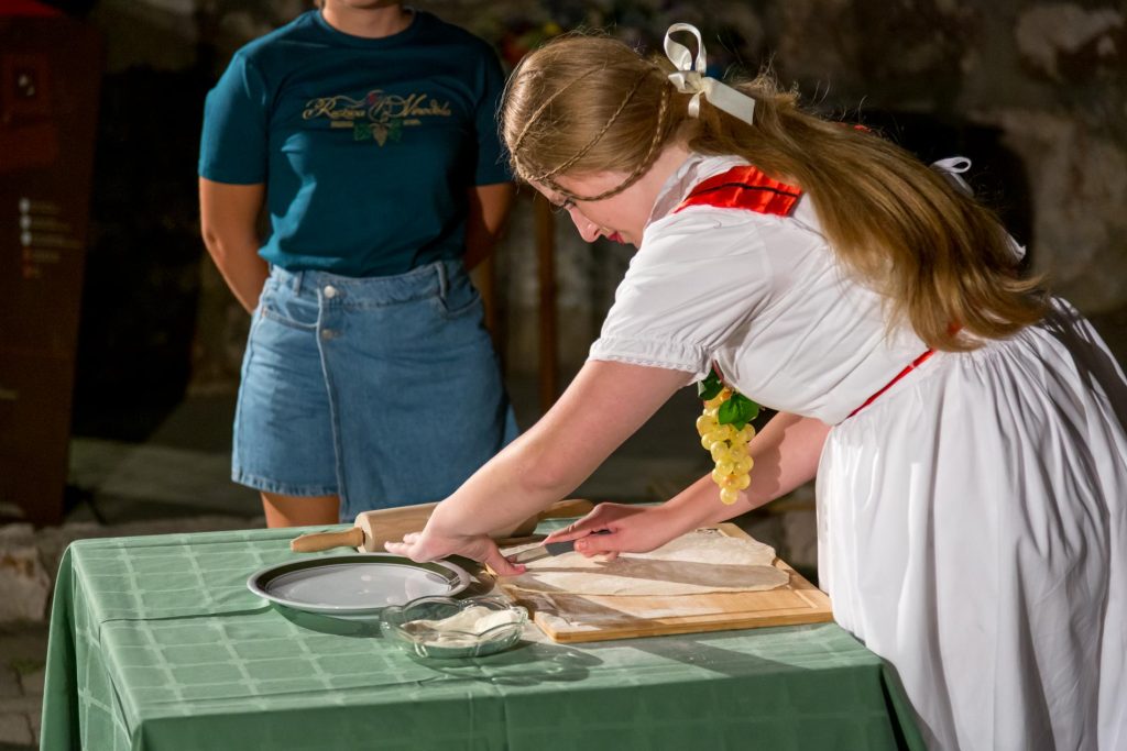 Woman in a white dress with red trim rolls dough on a wooden board at a green table while a man watches in the background.