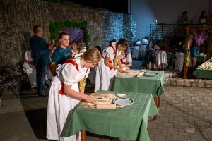 Women in white dresses with red sashes roll and cut dough on boards at a nighttime outdoor event on green-tablecloth tables.
