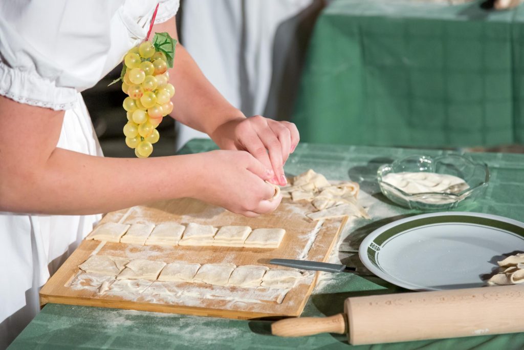 Person in white clothing shapes small pieces of dough on a flour-dusted wooden board; a rolling pin and plate sit nearby on a green tablecloth.