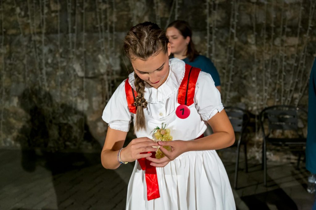 Young woman in a white dress with a red sash shows a grape skewer, wearing a pink badge with the number 7 in a dim setting