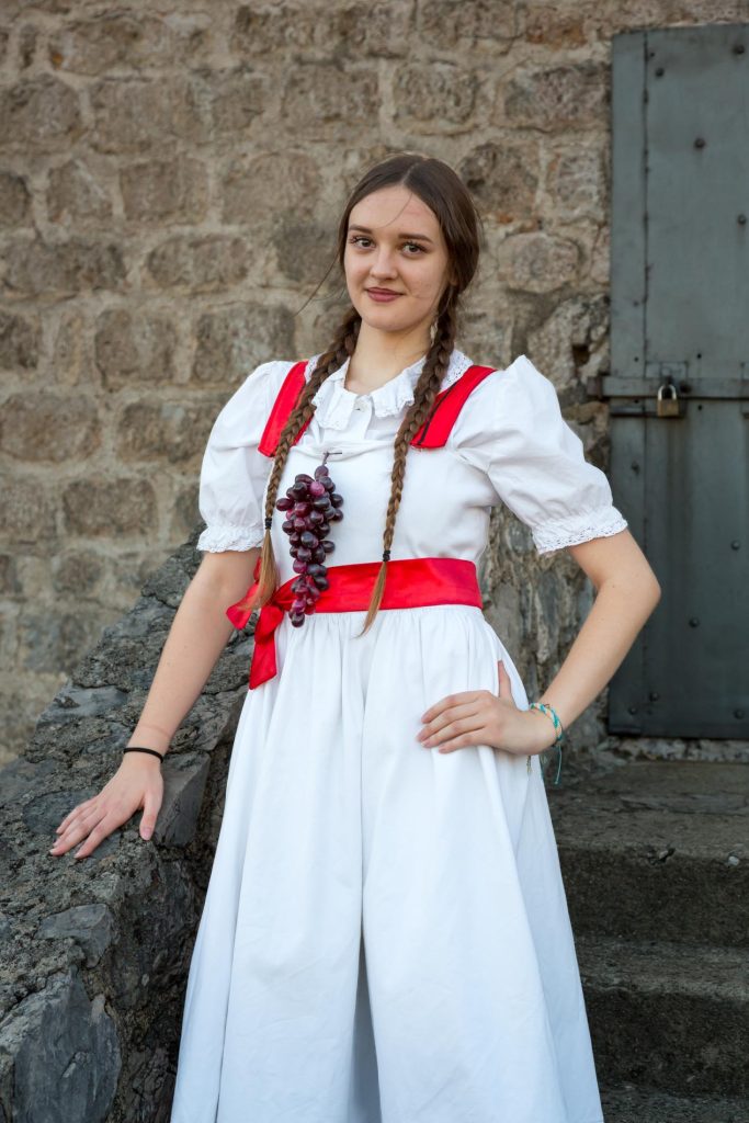 Young woman in a white dress with red sash and grape necklace, posing outdoors by a stone wall and door with braided hair.