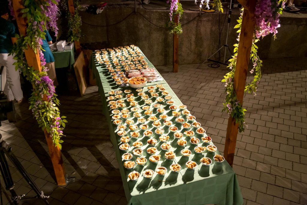 Table set for an event with dozens of small dessert-like cups arranged on a long green table under floral arches.