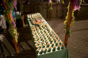 Table set for an event with dozens of small dessert-like cups arranged on a long green table under floral arches.