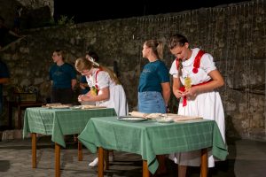 People in traditional outfits serving snacks at a night outdoor event with green tablecloths and a stone wall backdrop.