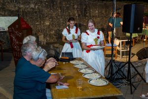 Two women in white dresses with red sashes serve plates of dumplings to seated guests at an outdoor event by a stone wall under string lights.