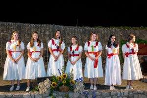 Group of young women in white dresses with red sashes and numbered badges, standing in a line on a stage with a flower basket in front during a night performance.