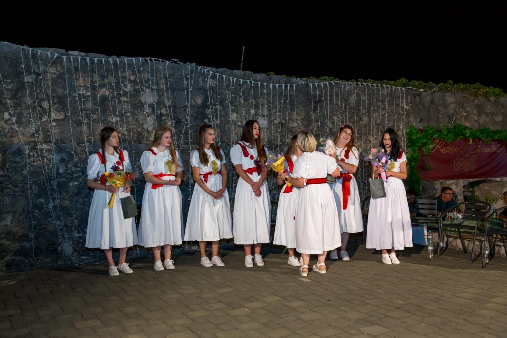 Group of young women in white dresses with red sashes standing in a line outdoors at night, holding bouquets.