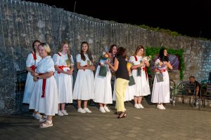 Group of young women in white dresses with red sashes receive flowers on a nighttime outdoor stage, beside a stone wall with string lights.