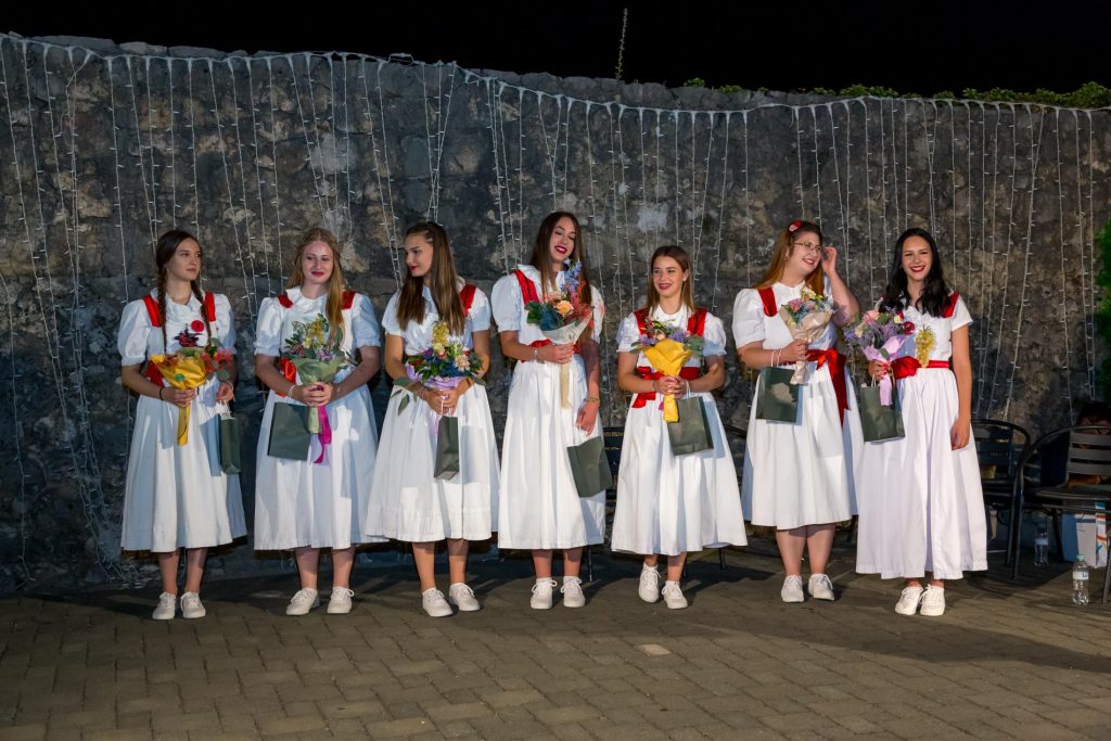 Seven women in white dresses with red sashes stand in a line outdoors at night, each holding bouquets and gift bags against a stone wall with string lights above.