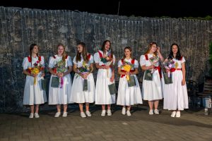 Seven women in white dresses with red sashes stand in a line outdoors at night, each holding bouquets and gift bags against a stone wall with string lights above.