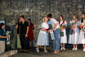 Two adults kiss on stage while others in dresses hold flowers and gifts against a stone wall backdrop.