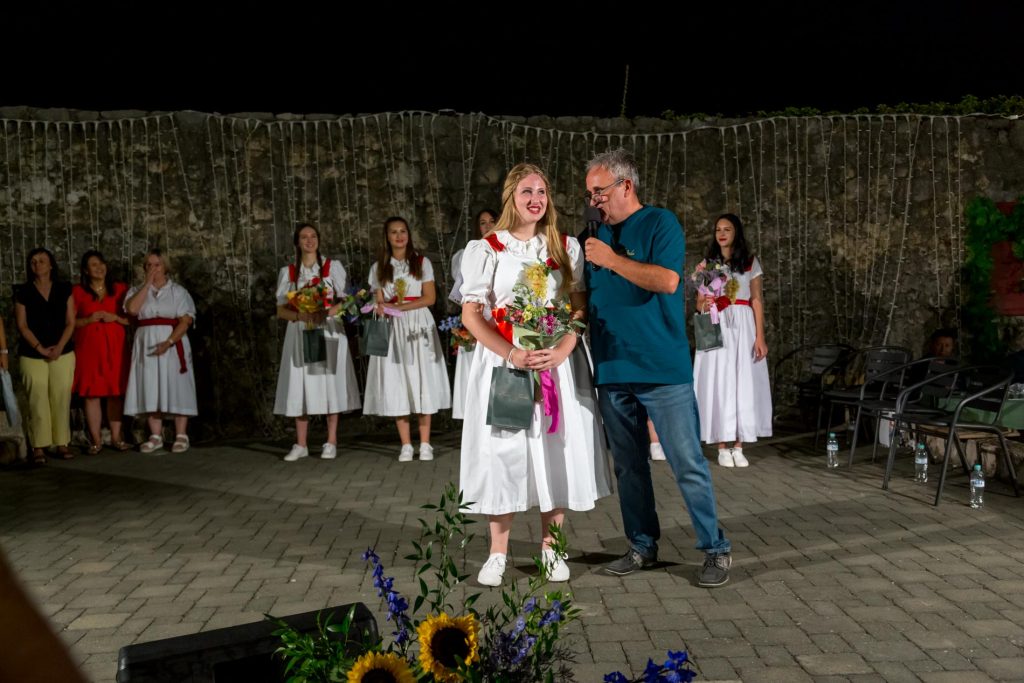Young woman in a white dress receiving flowers and a gift bag on stage, man with a microphone beside her at a nighttime ceremony.