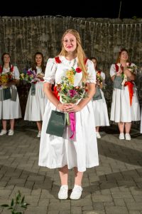 Center-front woman in a white dress holds a bouquet and a gift bag at a nighttime outdoor ceremony, with others in similar dresses in the background.